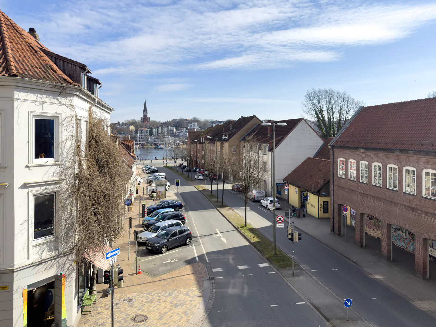 Street view of a European town with cars parked along the side, buildings with red tile roofs, and a church steeple in the distance.