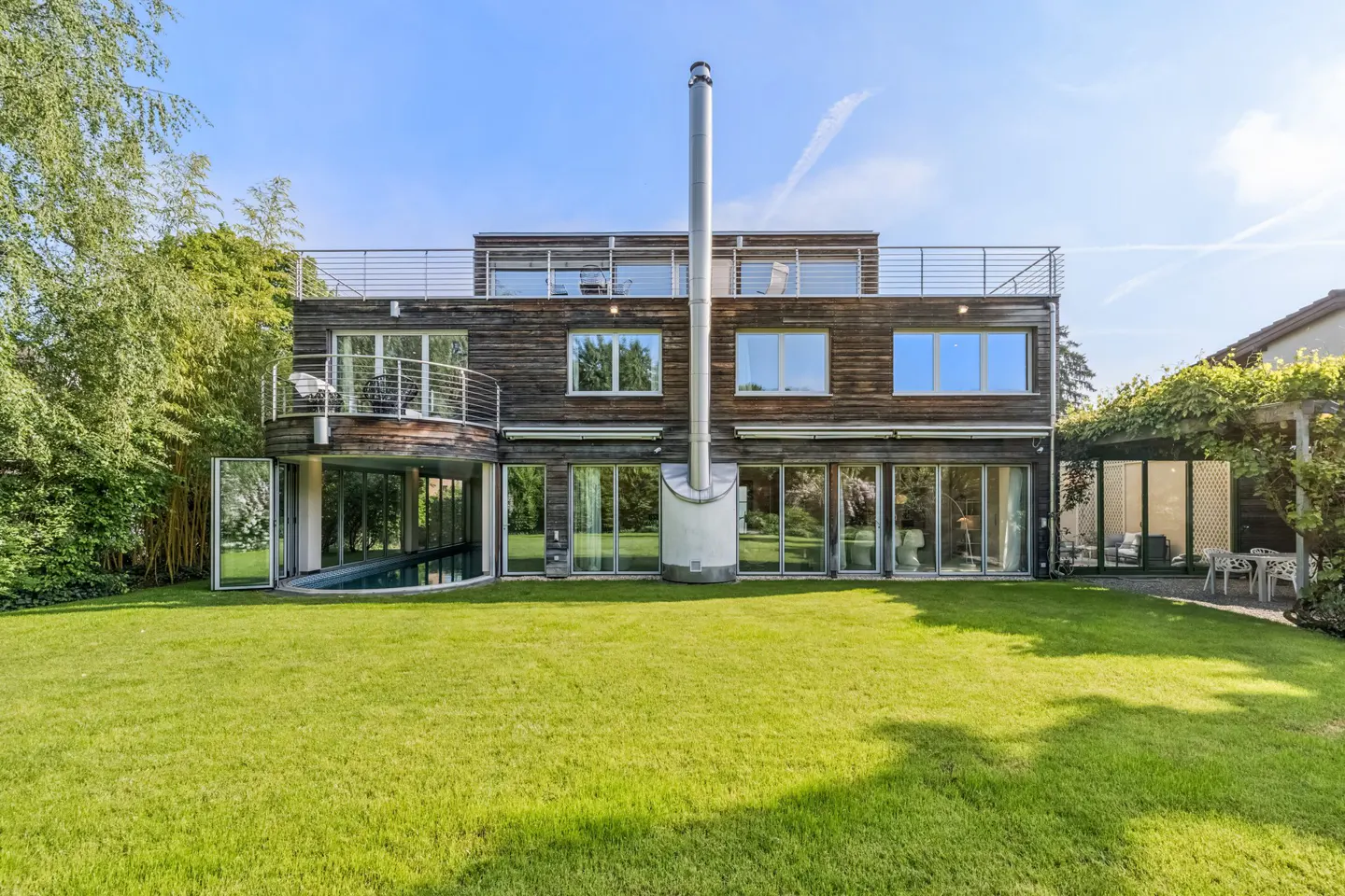 Modern three-story house with wood siding, large windows, a tall chimney, and a green lawn. A pool is visible under a balcony.