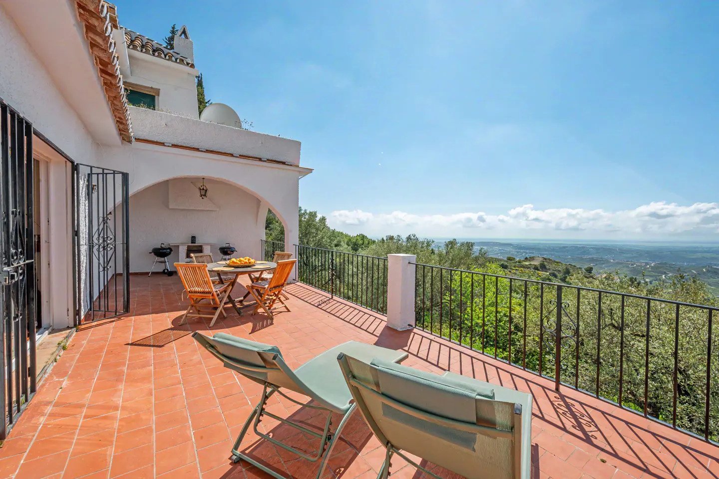 Terracotta-tiled patio with two lounge chairs, a table with fruit, and a grill. Black railing overlooks a green, hilly landscape under a blue sky.