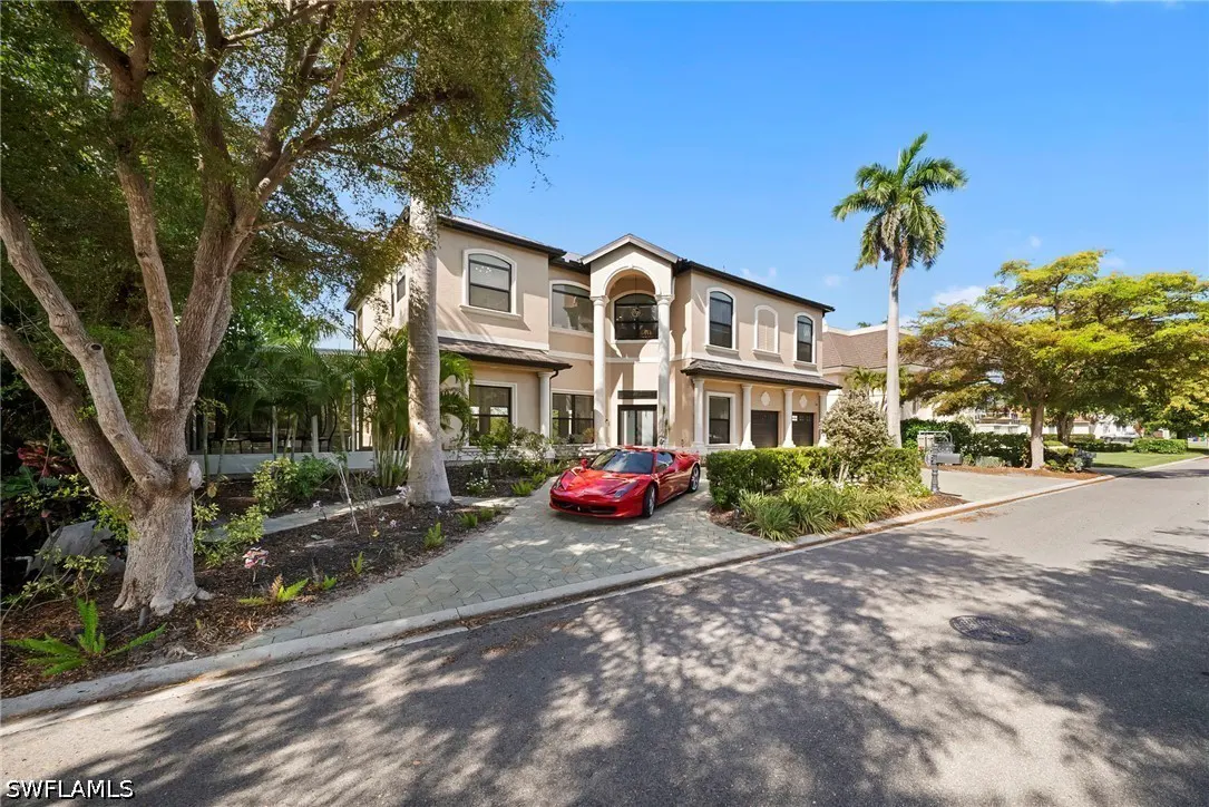 Two-story beige house with a red sports car parked in the driveway, surrounded by trees and greenery.