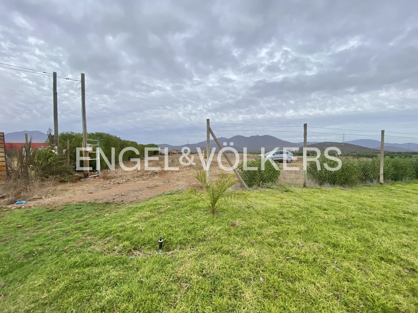 A grassy foreground leads to a fenced, dry dirt lot under a cloudy sky with distant mountains in the background.