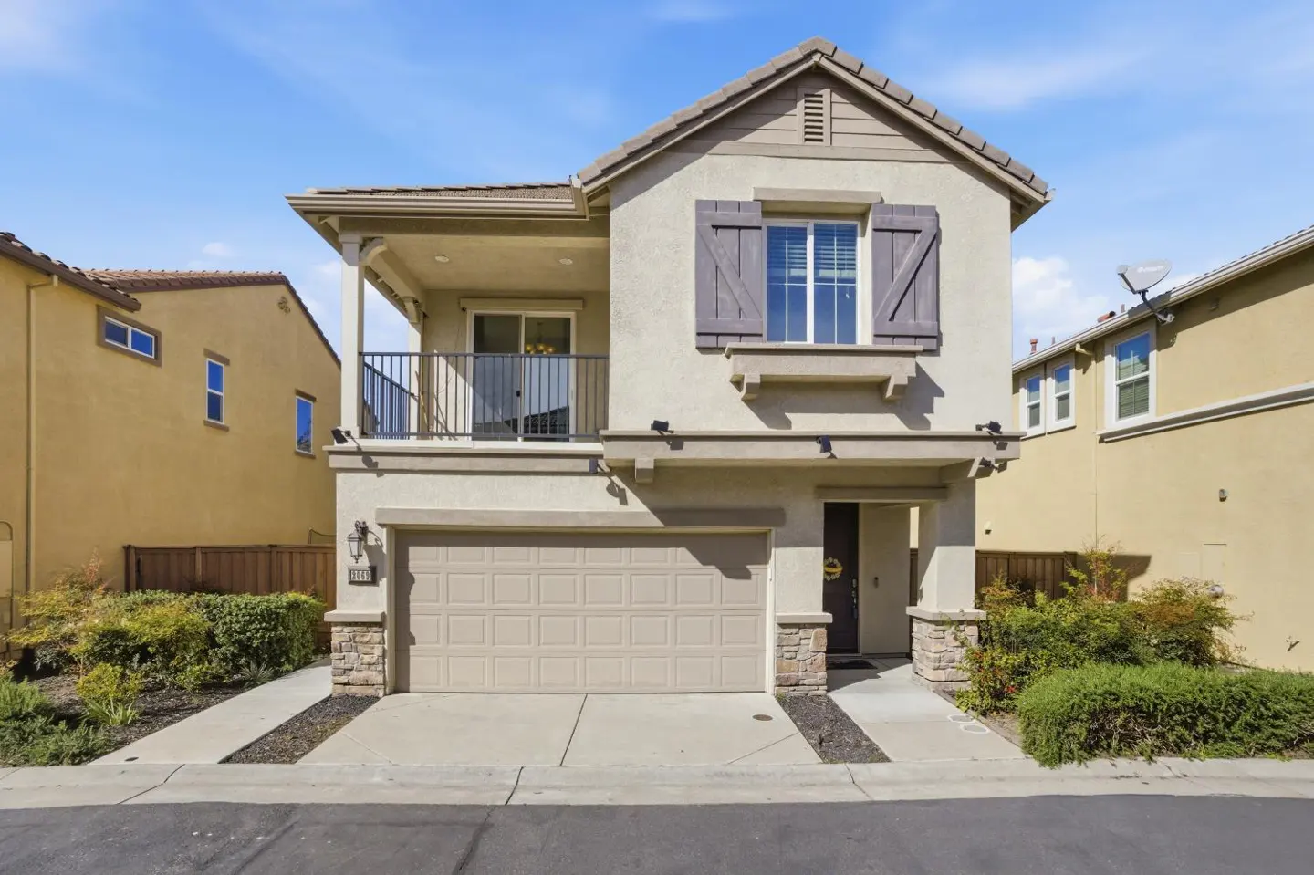 Two-story beige house with a balcony, gray shutters, and a tan garage door under a blue sky.