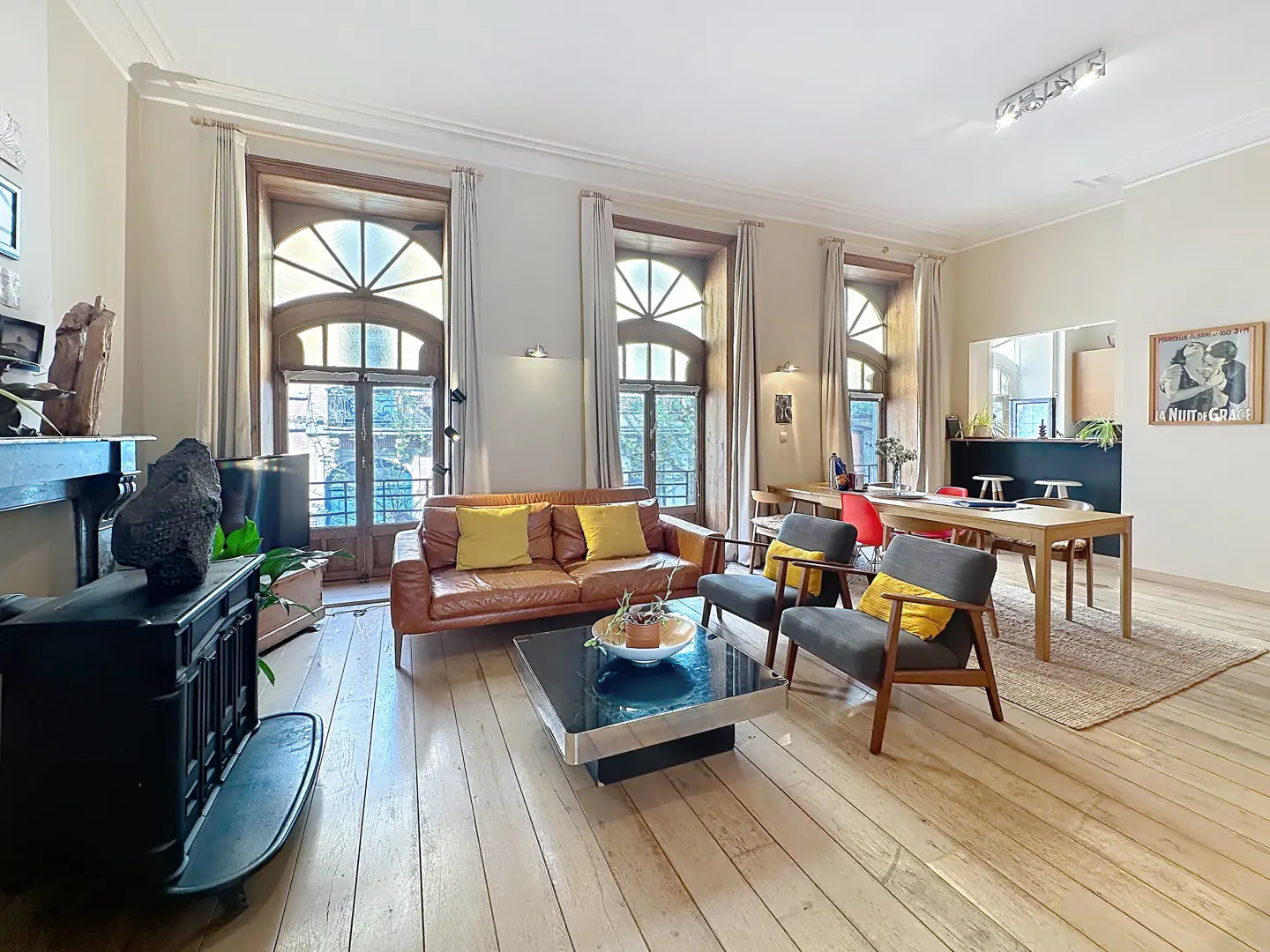 Bright living room with wood floors, a brown leather sofa with yellow pillows, and arched windows with beige curtains.