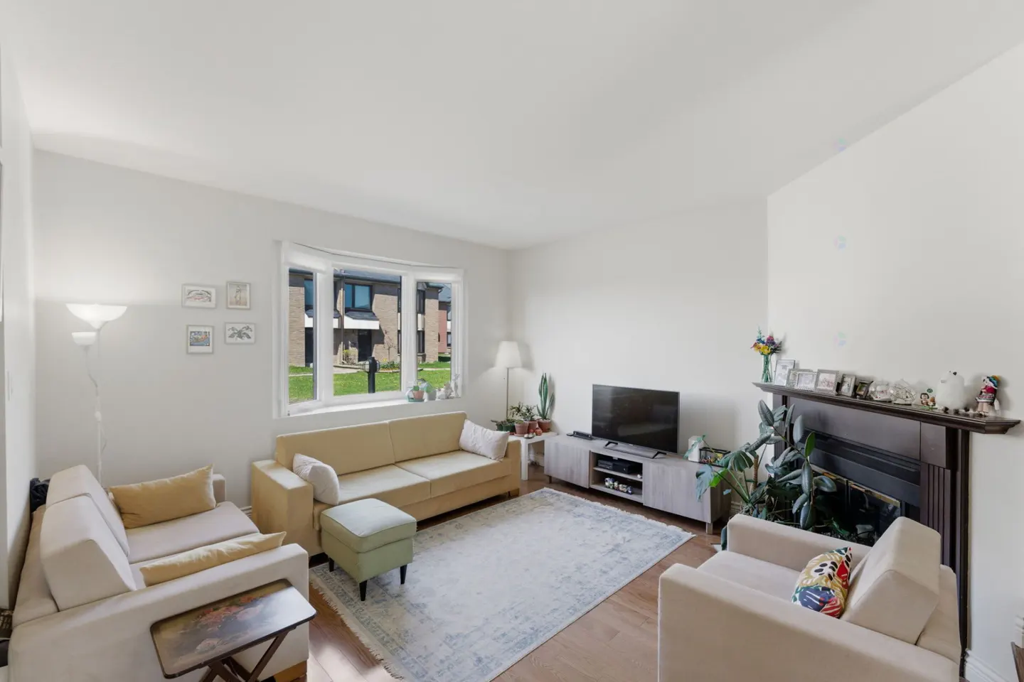 Bright living room with beige sofas, a fireplace, and a large window overlooking a green lawn and houses.