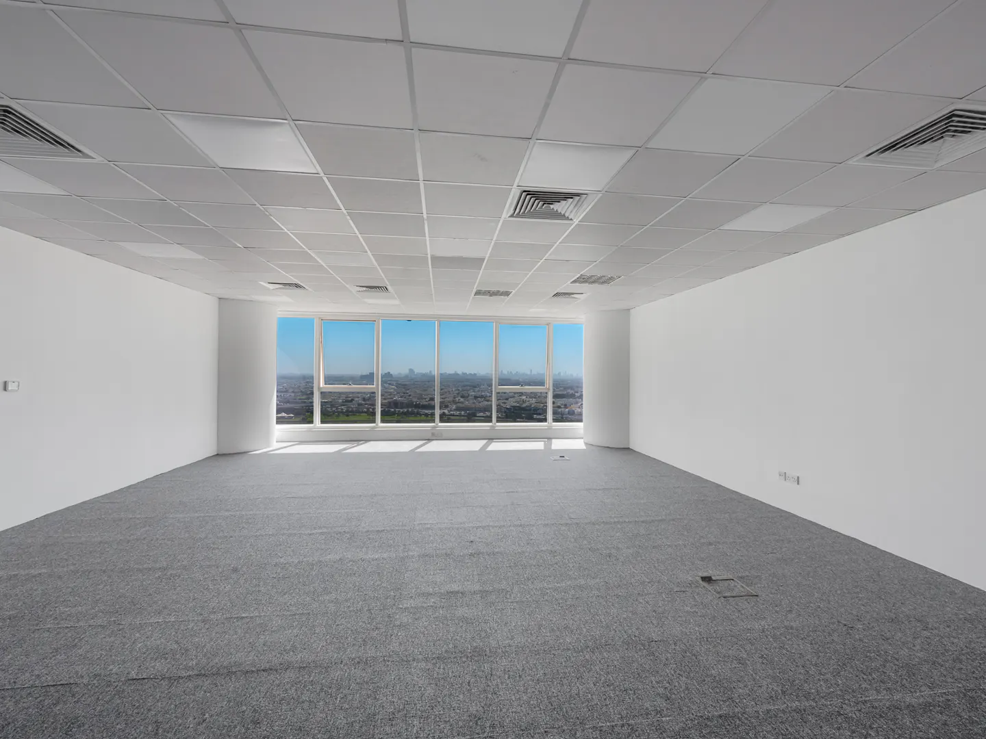 Empty office space with gray carpet, white walls, and a large window showing a city skyline under a blue sky.