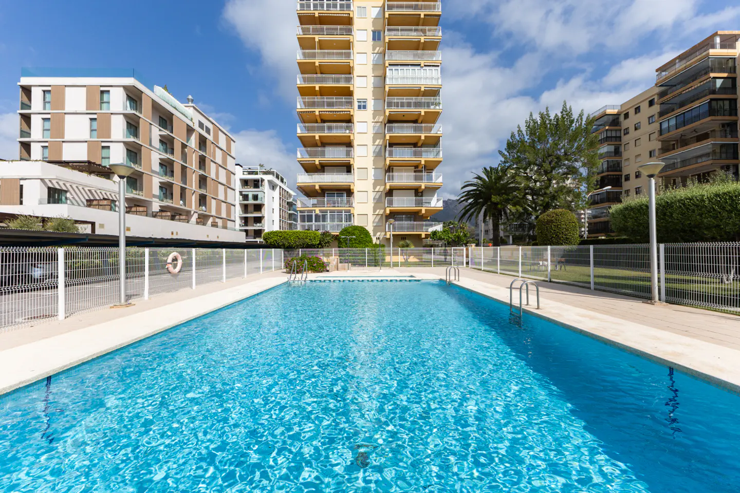 Outdoor pool with clear blue water, surrounded by white fence and buildings under a blue sky.