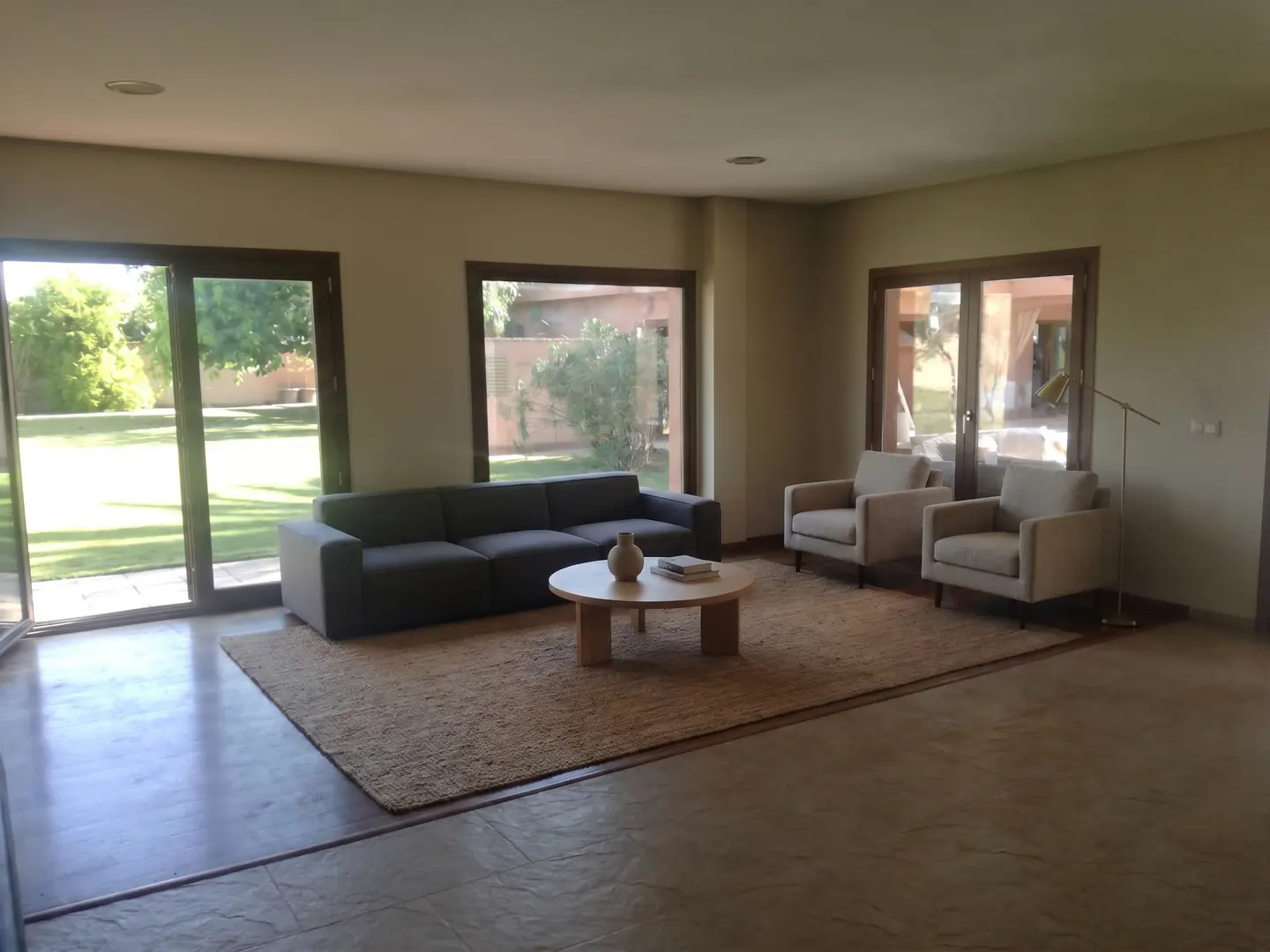 Living room with a gray sofa, two armchairs, and a round coffee table on a jute rug. Large windows overlook a green lawn.