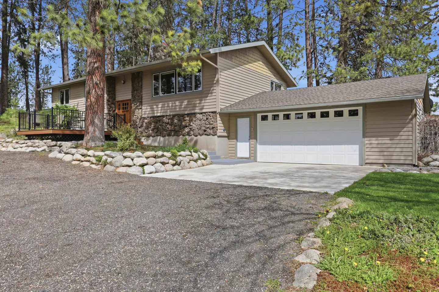 Tan two-story house with stone accents, a deck, and a white garage door, surrounded by trees.