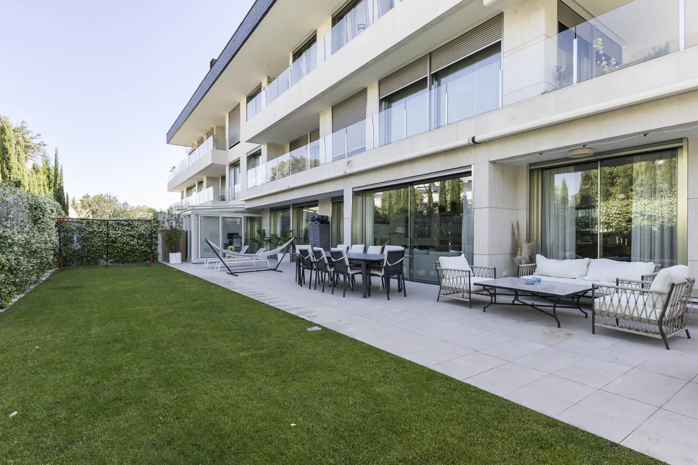 A modern building with a patio, lawn, and outdoor furniture. A hammock is set up near a dining table and chairs. A white sofa and chairs are on the patio.