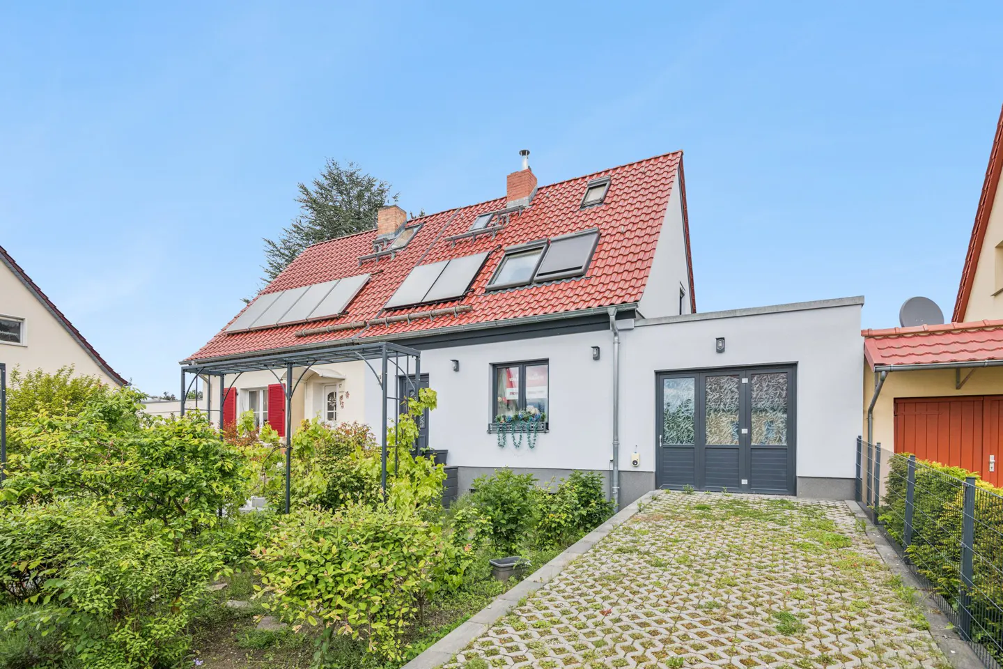 A white house with a red tile roof and solar panels, a garden, and a paved driveway.