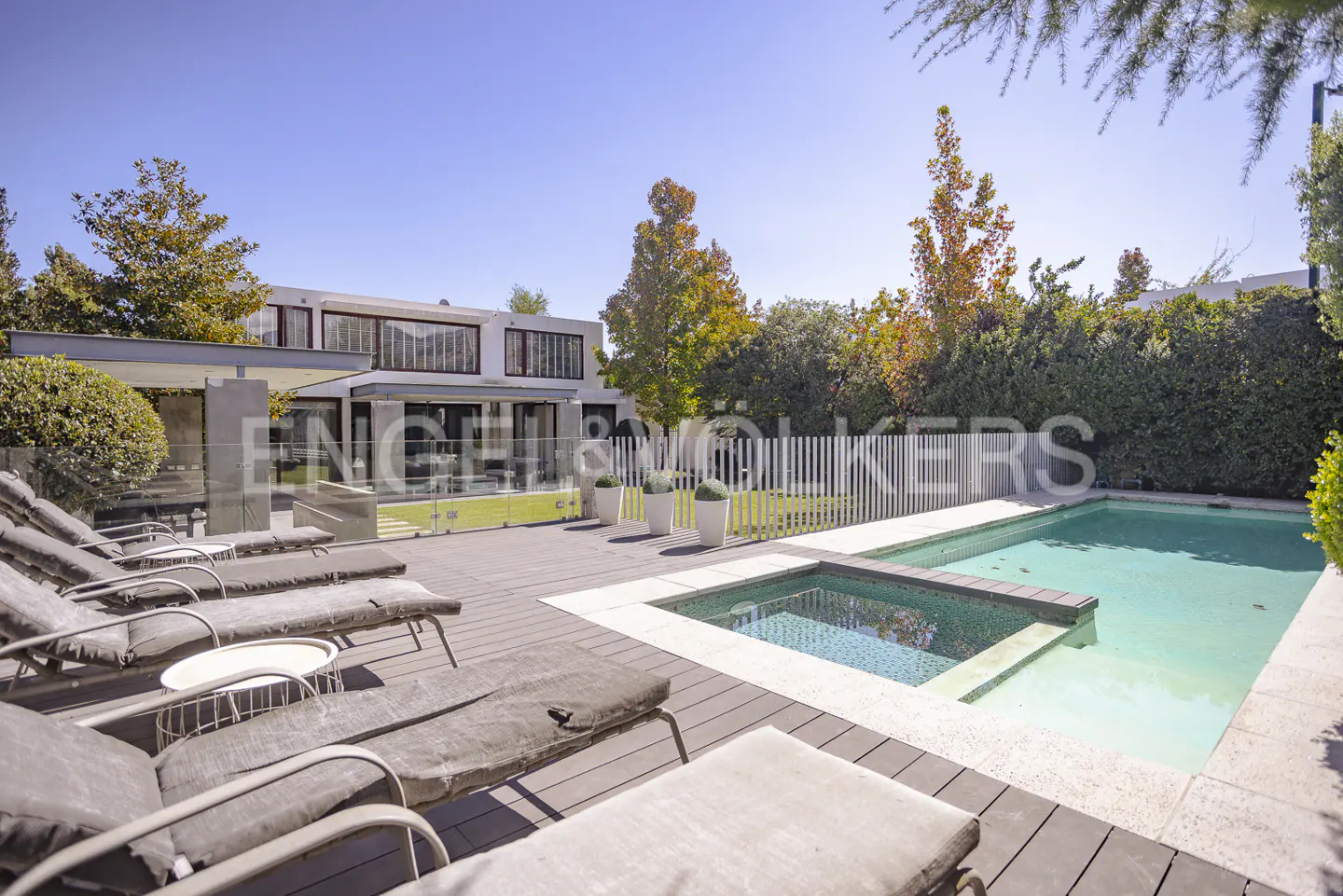 Modern home exterior with a pool and lounge chairs on a wooden deck under a clear blue sky.