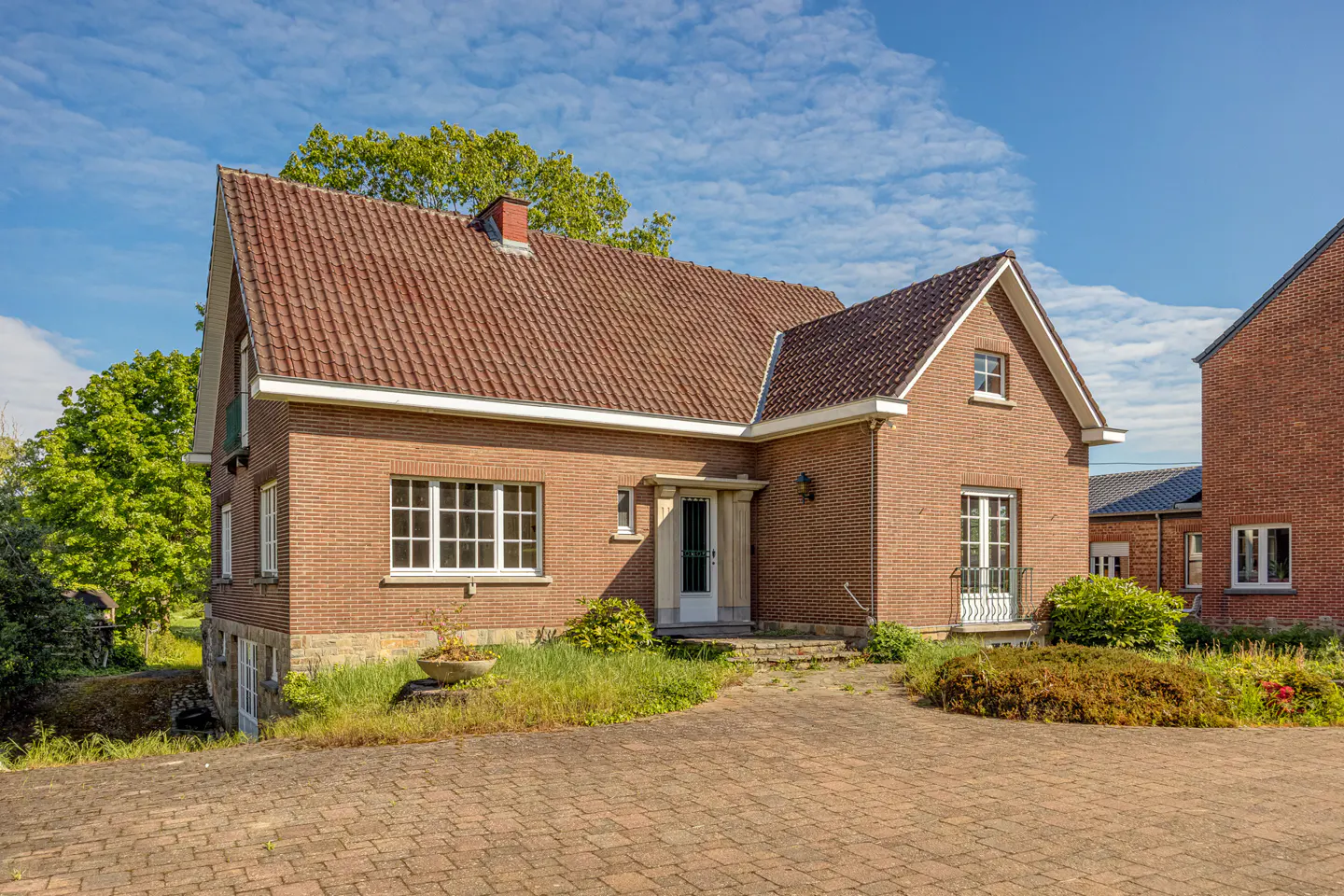 Two-story brick house with a brown tiled roof, white window frames, and a paved driveway under a blue sky.