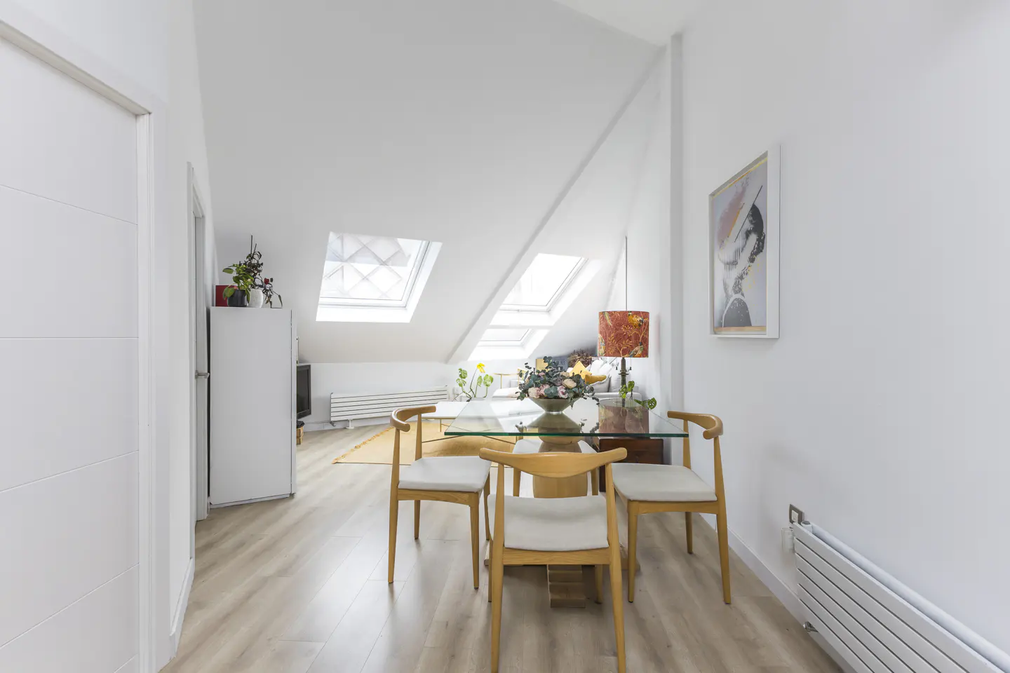 Bright, modern dining area with a glass table, four wooden chairs, and skylights in the sloped ceiling. White walls and light wood floors.