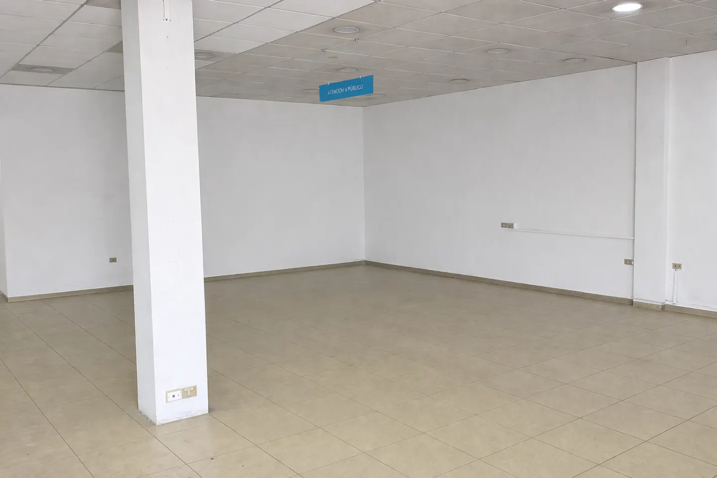 Empty commercial space with white walls, beige tiled floor, and a large white support column. A blue sign hangs from the ceiling.