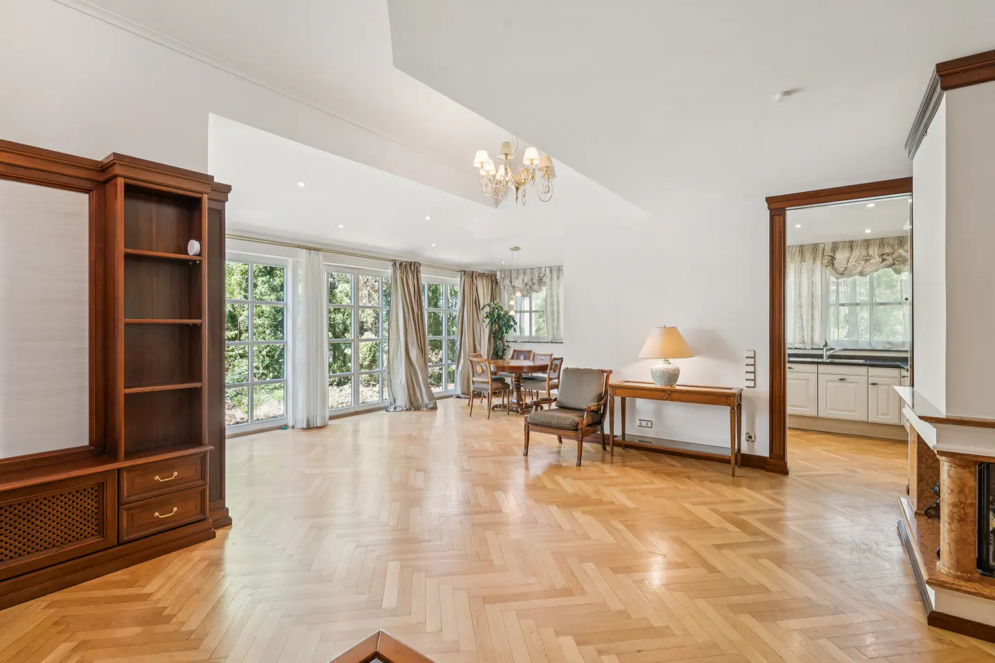 Bright living room with herringbone wood floors, white walls, and large windows. A dining table, chairs, and a wooden bookcase are visible.