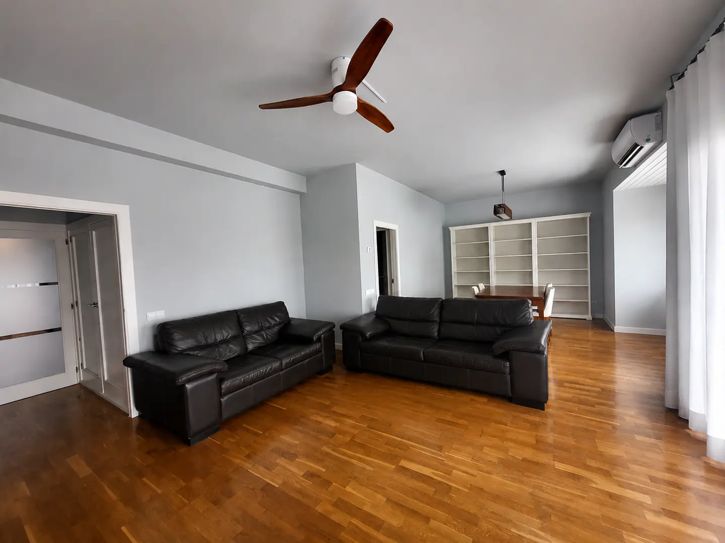 Spacious living room with two black leather sofas, hardwood floors, a wooden ceiling fan, and a built-in white bookshelf.