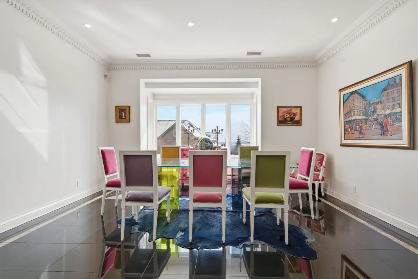 Dining room with a glass table, colorful chairs, and a blue rug on a black tile floor. A large window shows a snowy landscape.