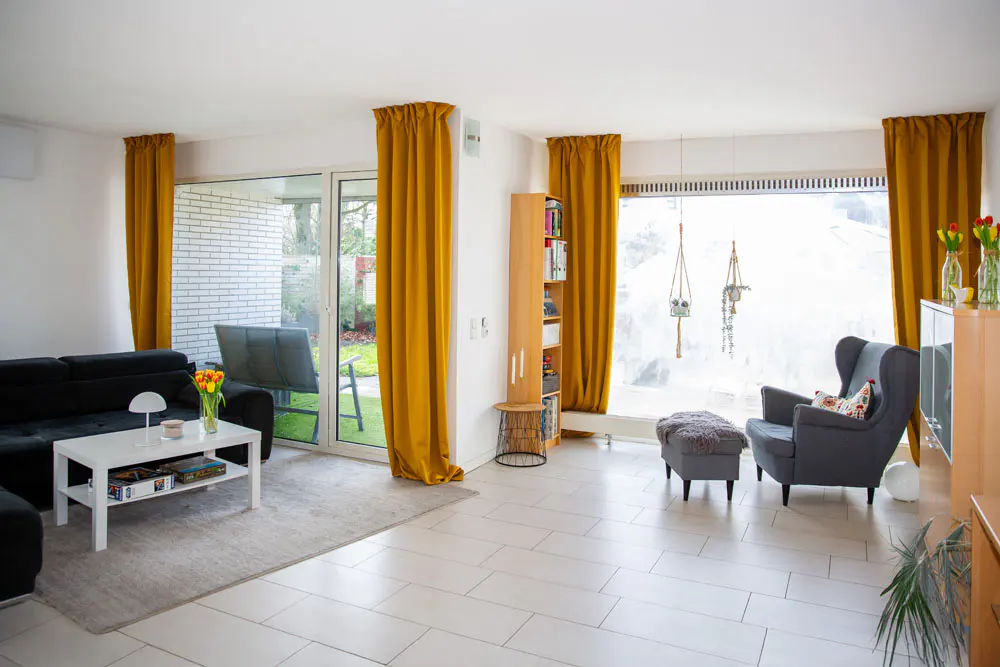 Bright living room with white tile floors, black sofa, gray armchair, and yellow curtains framing windows and a sliding glass door.
