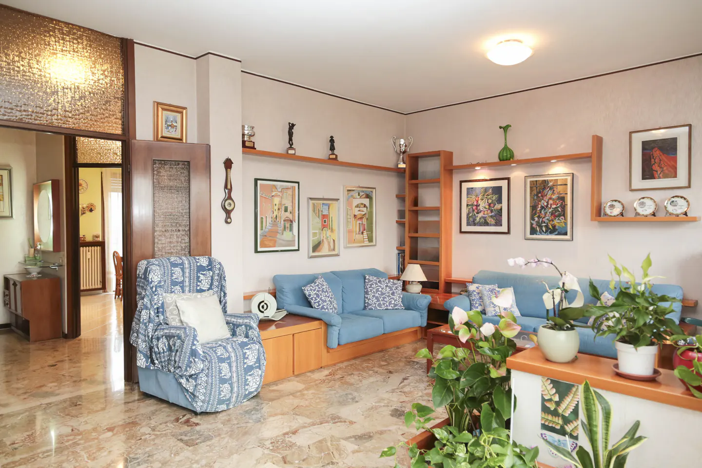 A living room with blue sofas, wooden shelves, and marble floors. Artwork and plants decorate the space.