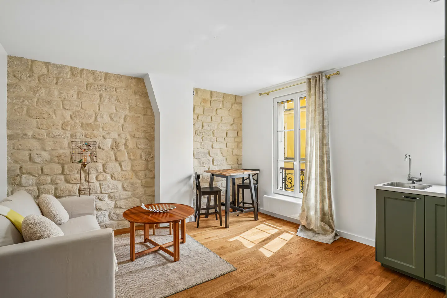 Bright apartment interior with stone walls, hardwood floors, and a window with patterned curtains. A white sofa, wooden tables, and a small kitchen area are visible.
