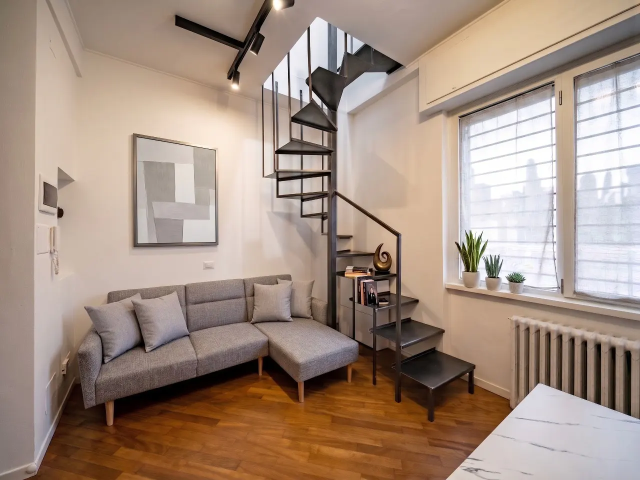 Living room with a gray sofa, wood floors, and a black spiral staircase. A modern art piece hangs on the wall. Natural light streams through the window.