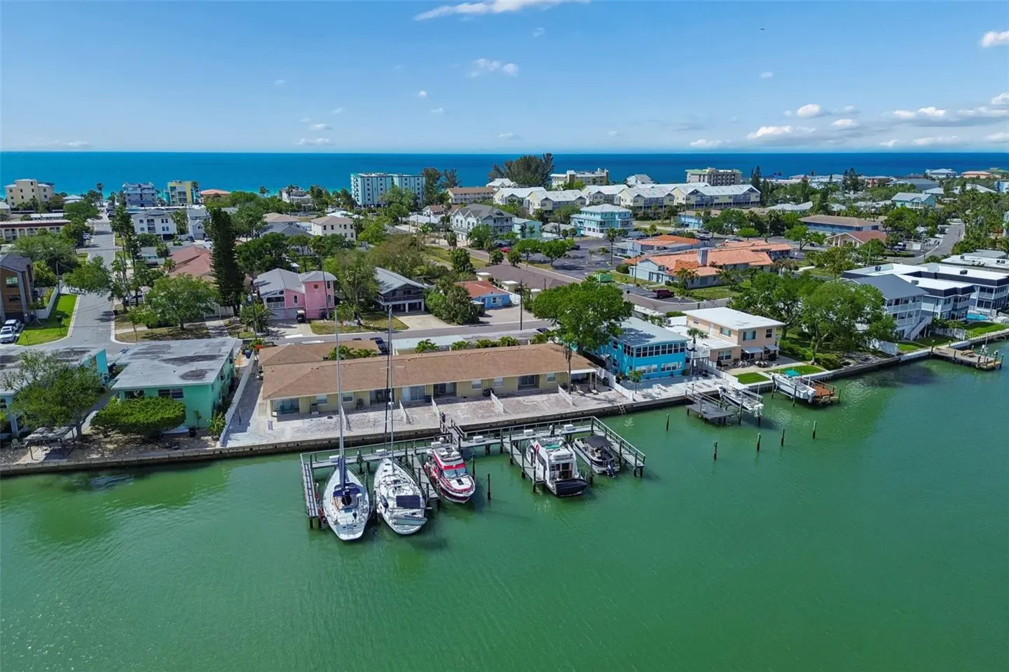 Aerial view of waterfront homes with boats docked on a sunny day. Blue ocean and sky in the background.