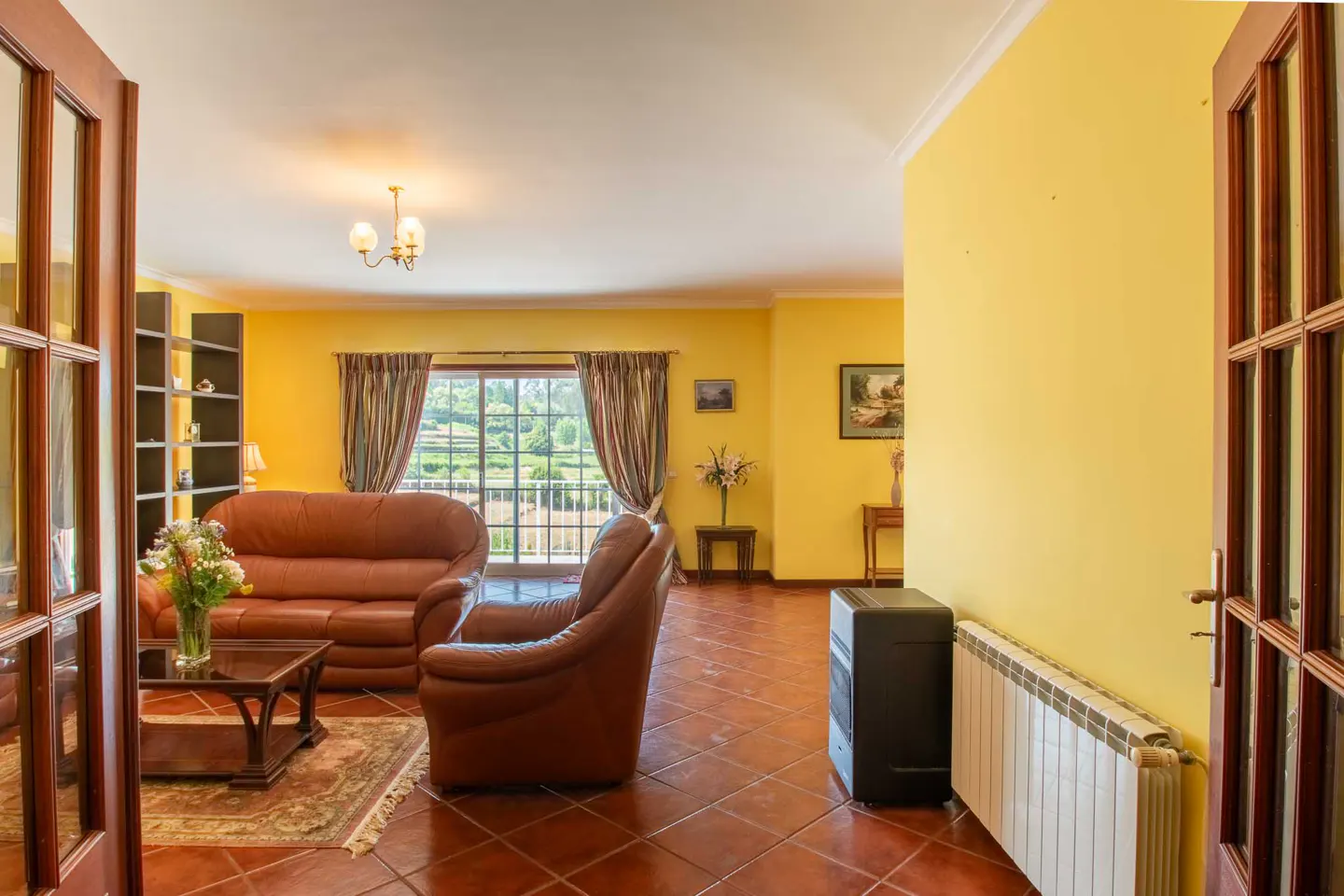 Living room with yellow walls, brown leather sofa and armchair, and a large window with a view of the garden.