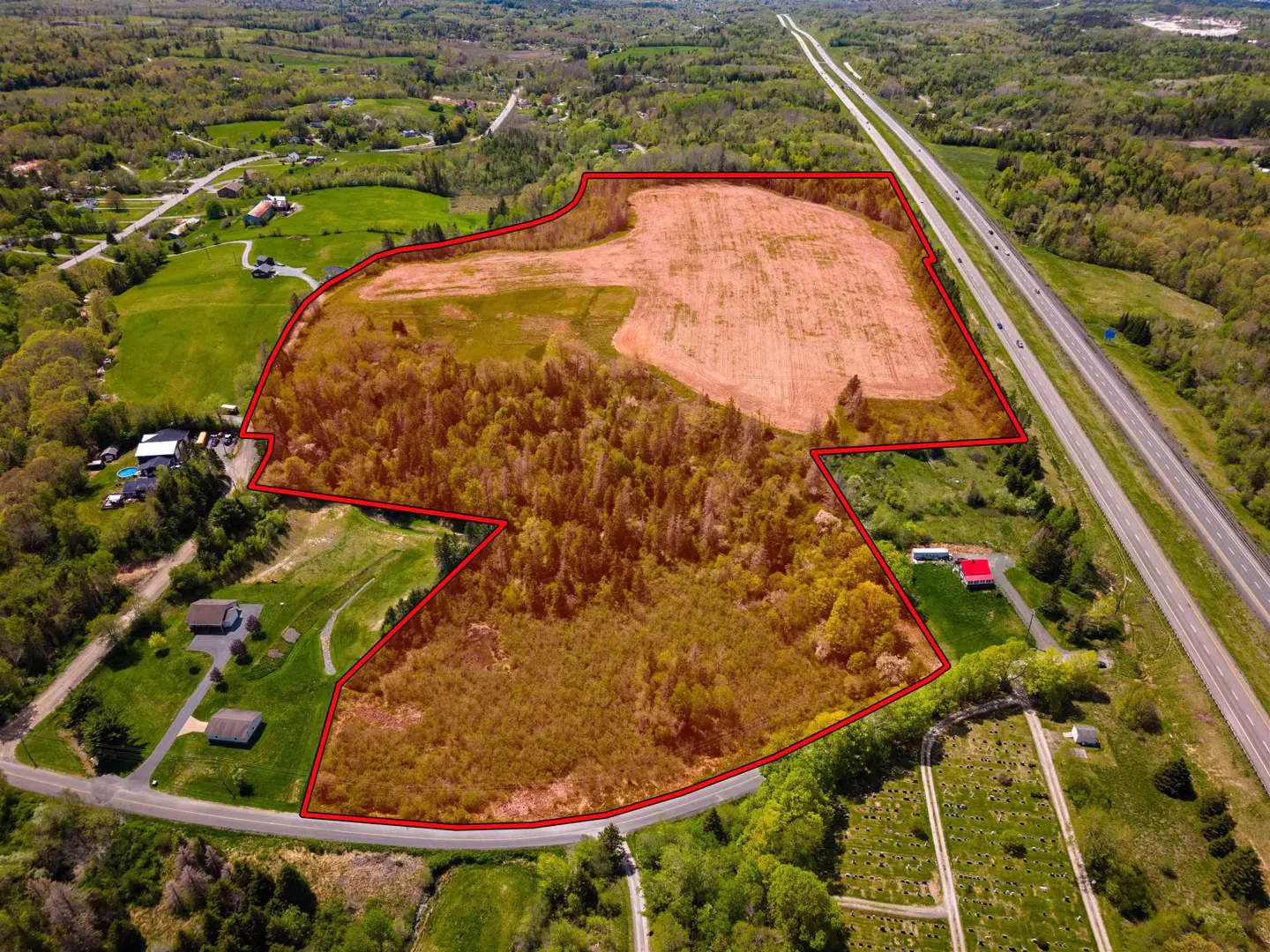 Aerial view of a large, partially wooded plot of land outlined in red, situated next to a highway and rural homes.