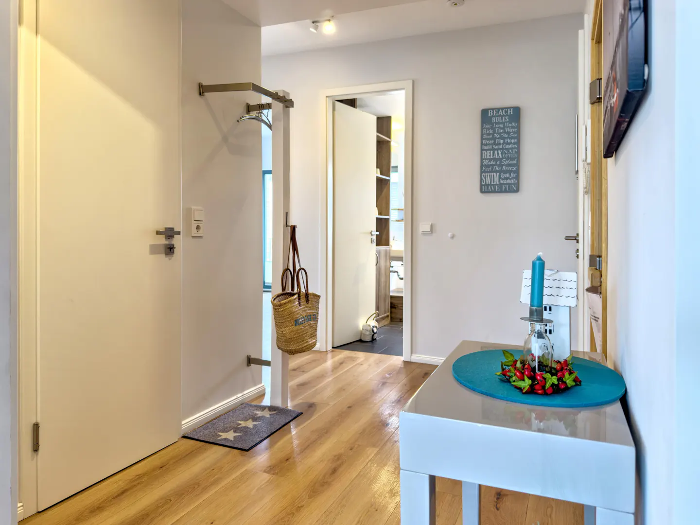 Hallway with wood floors, white walls, and a white table with a blue candle and red berries. A straw bag hangs near a door.