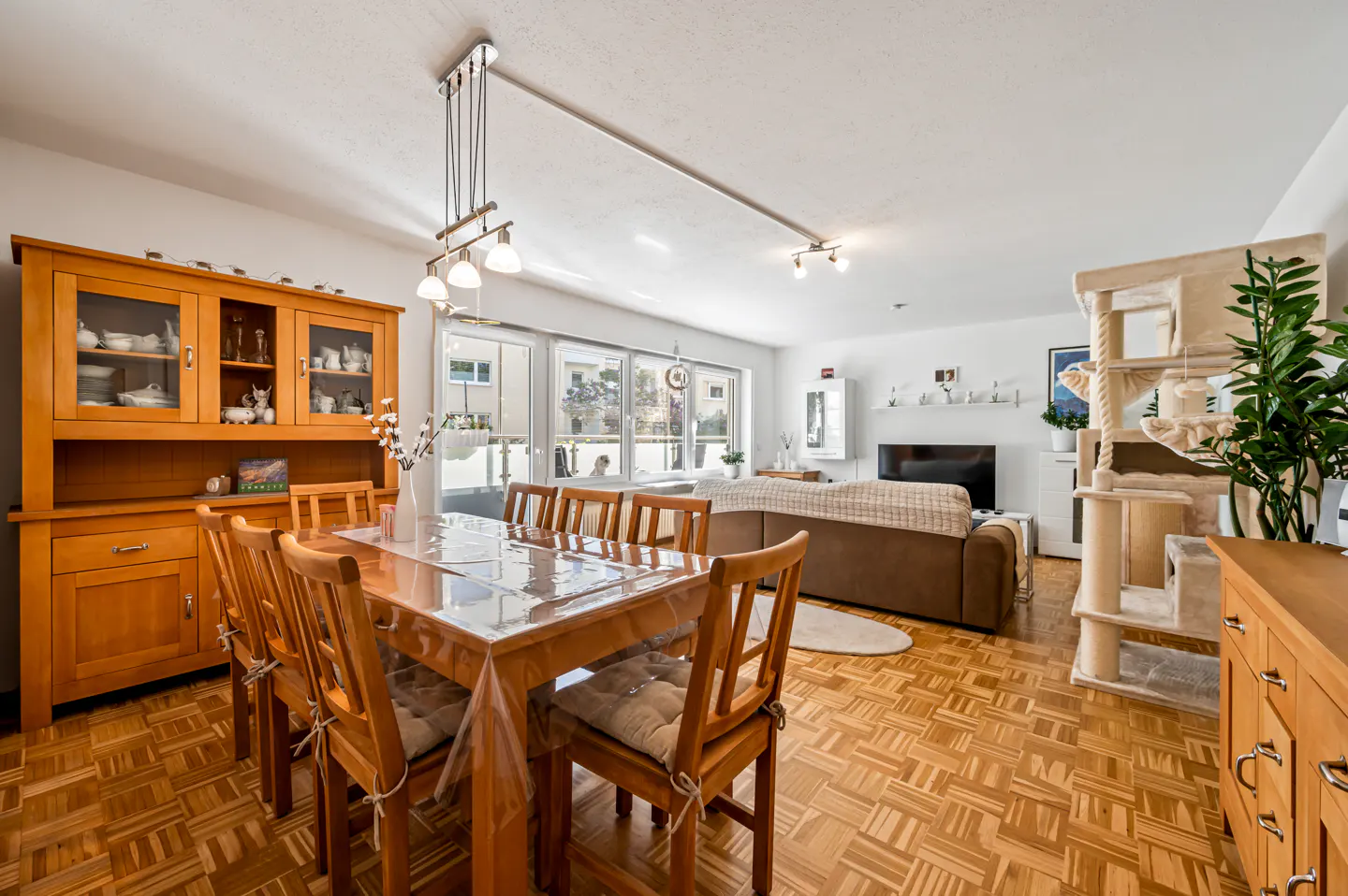 Bright living and dining area with wood parquet flooring. A wooden dining table and hutch are visible, along with a brown sofa and cat tree.
