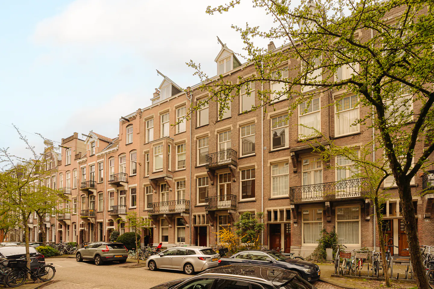 Street view of row houses in Amsterdam. Cars and bikes line the street. Trees with green leaves are in the foreground.