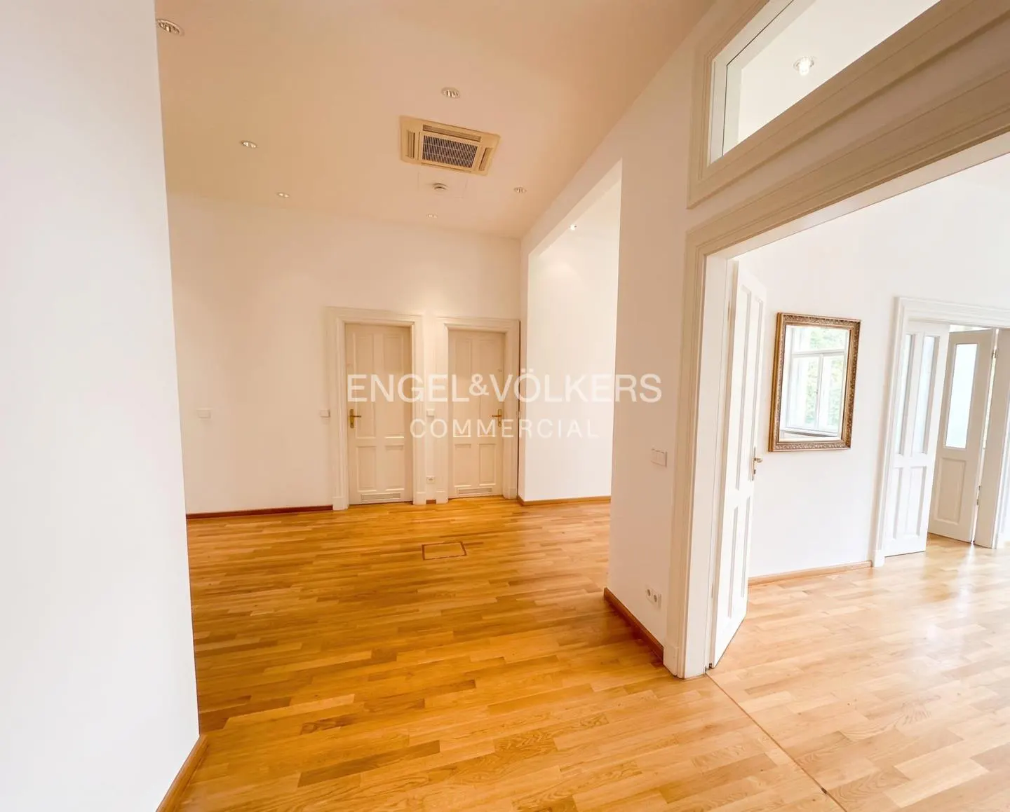 Bright, empty office space with light wood floors, white walls, multiple doorways, and a gold-framed mirror.