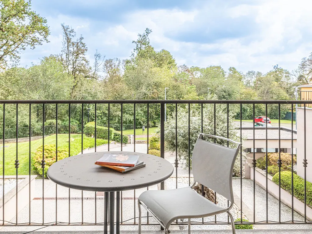 Balcony view with a gray chair and table holding books. Black railing overlooks green trees and a cloudy sky.