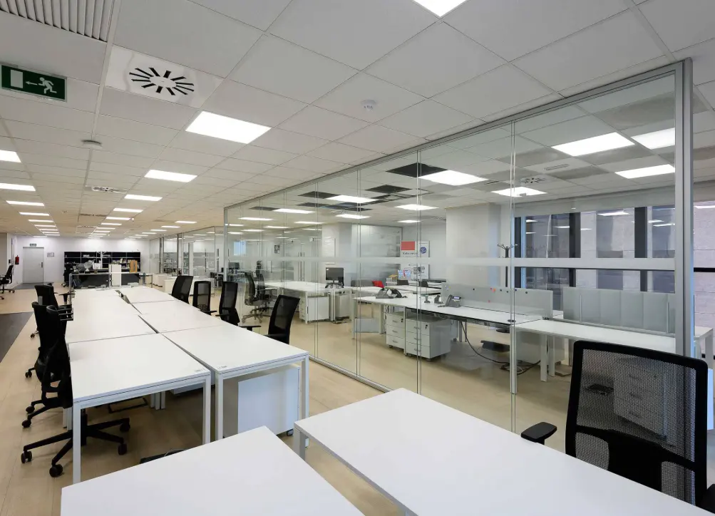 Bright, modern office space with white desks, black chairs, and glass-walled offices under a white, tiled ceiling with square lighting panels.