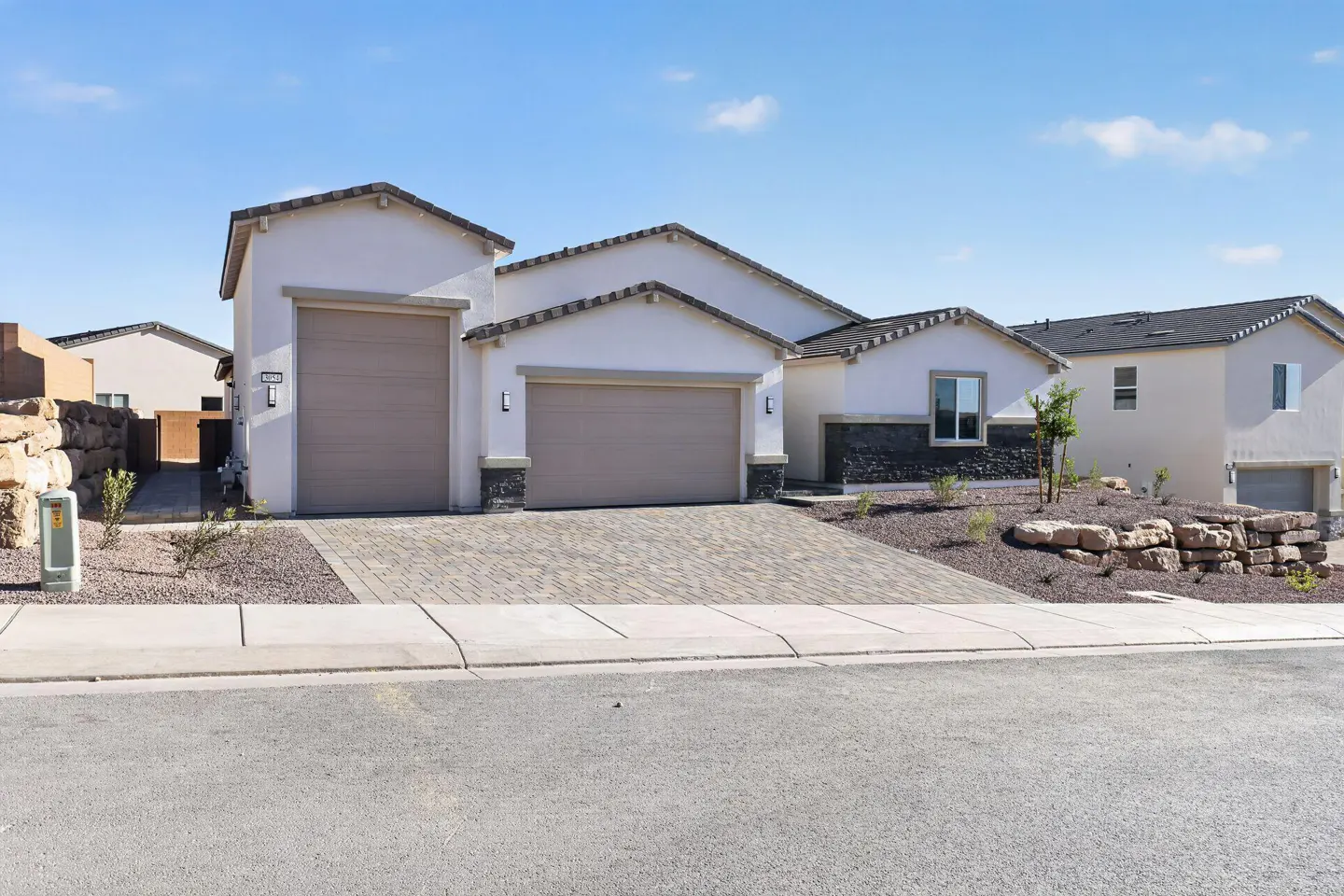 A modern, single-story stucco house with two garage doors and a brick driveway under a blue sky.