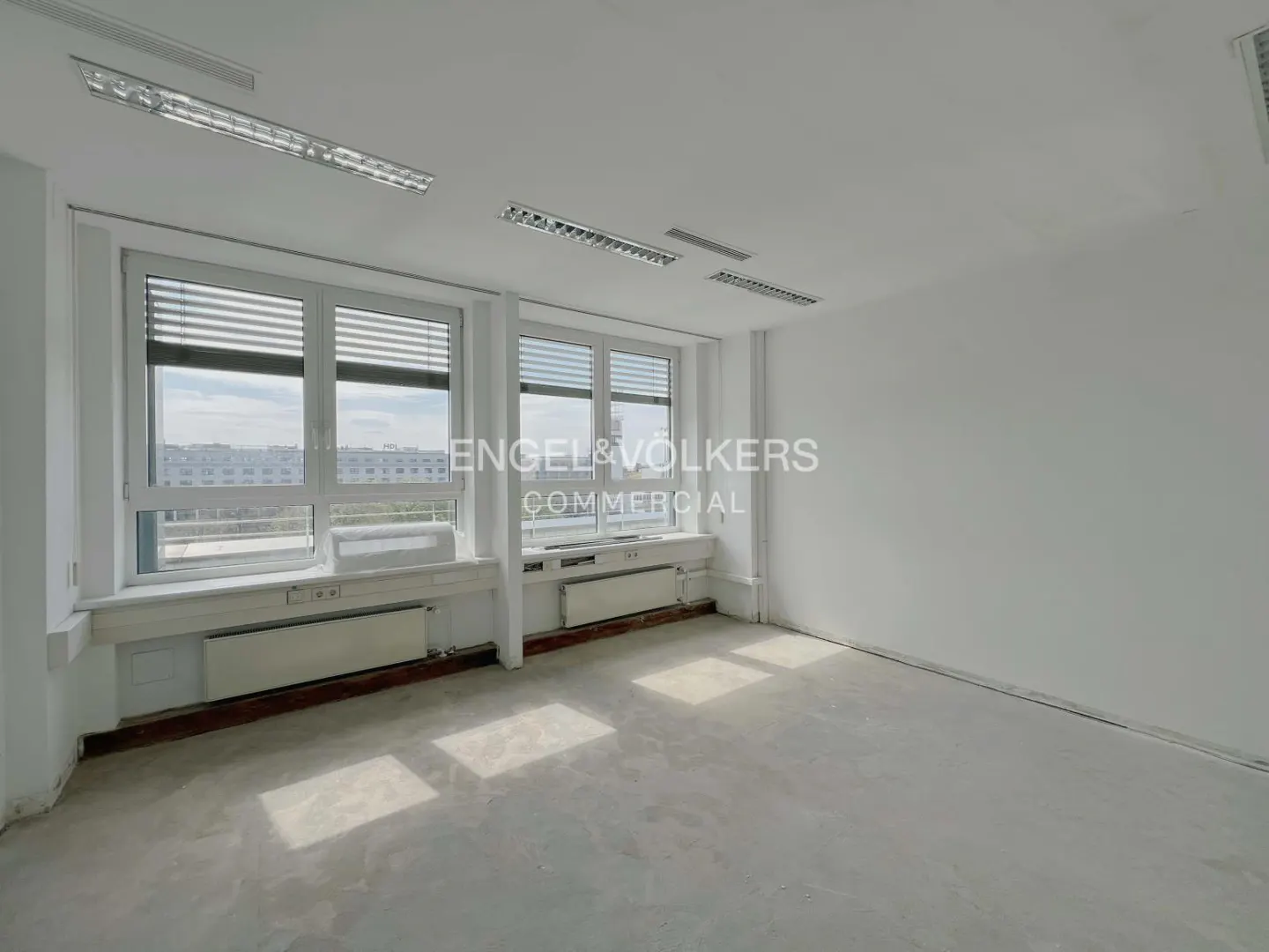 Bright, empty office space with white walls, gray floor, and large windows with blinds overlooking a cityscape.