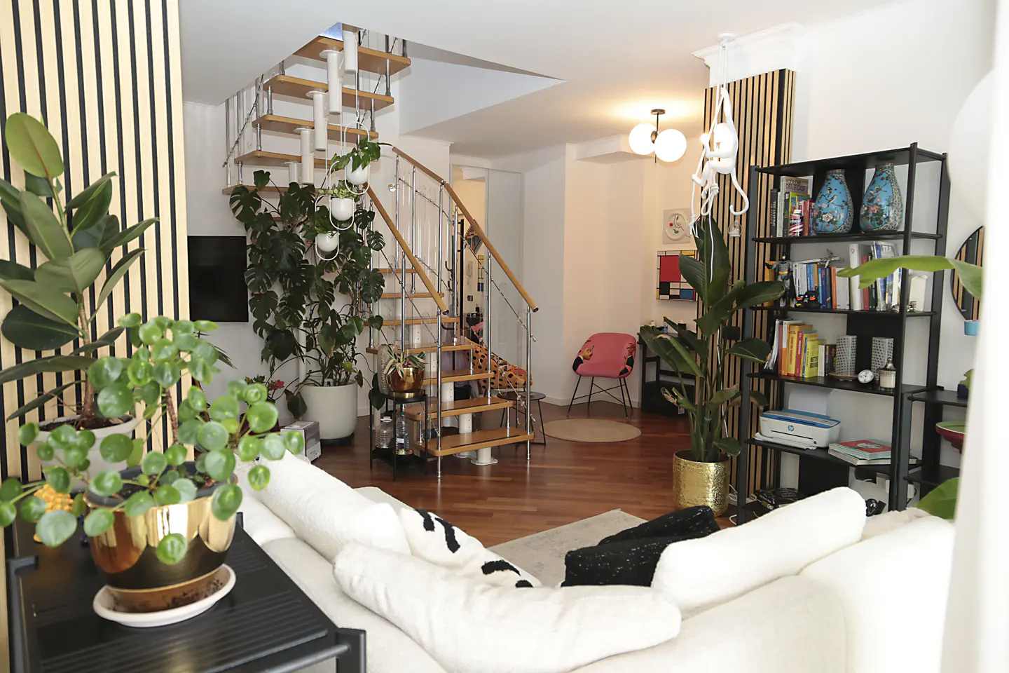 A bright living room with a wooden staircase, plants, a white sofa, and a black bookshelf.