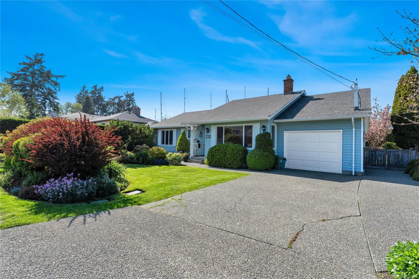 A light blue single-story house with a white garage door, green lawn, and a concrete driveway under a bright blue sky.