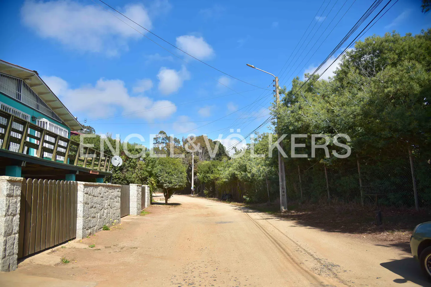 A dirt road leads past a turquoise house with a wooden fence and stone pillars, under a blue sky with scattered clouds.