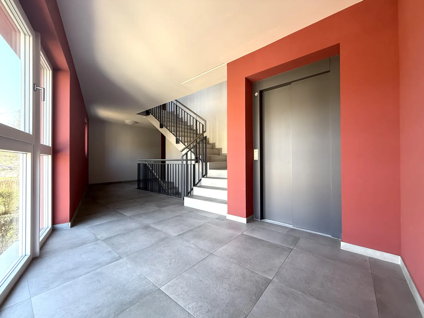 A building lobby with gray tile floors, a staircase with black railings, and a gray elevator with a red wall.