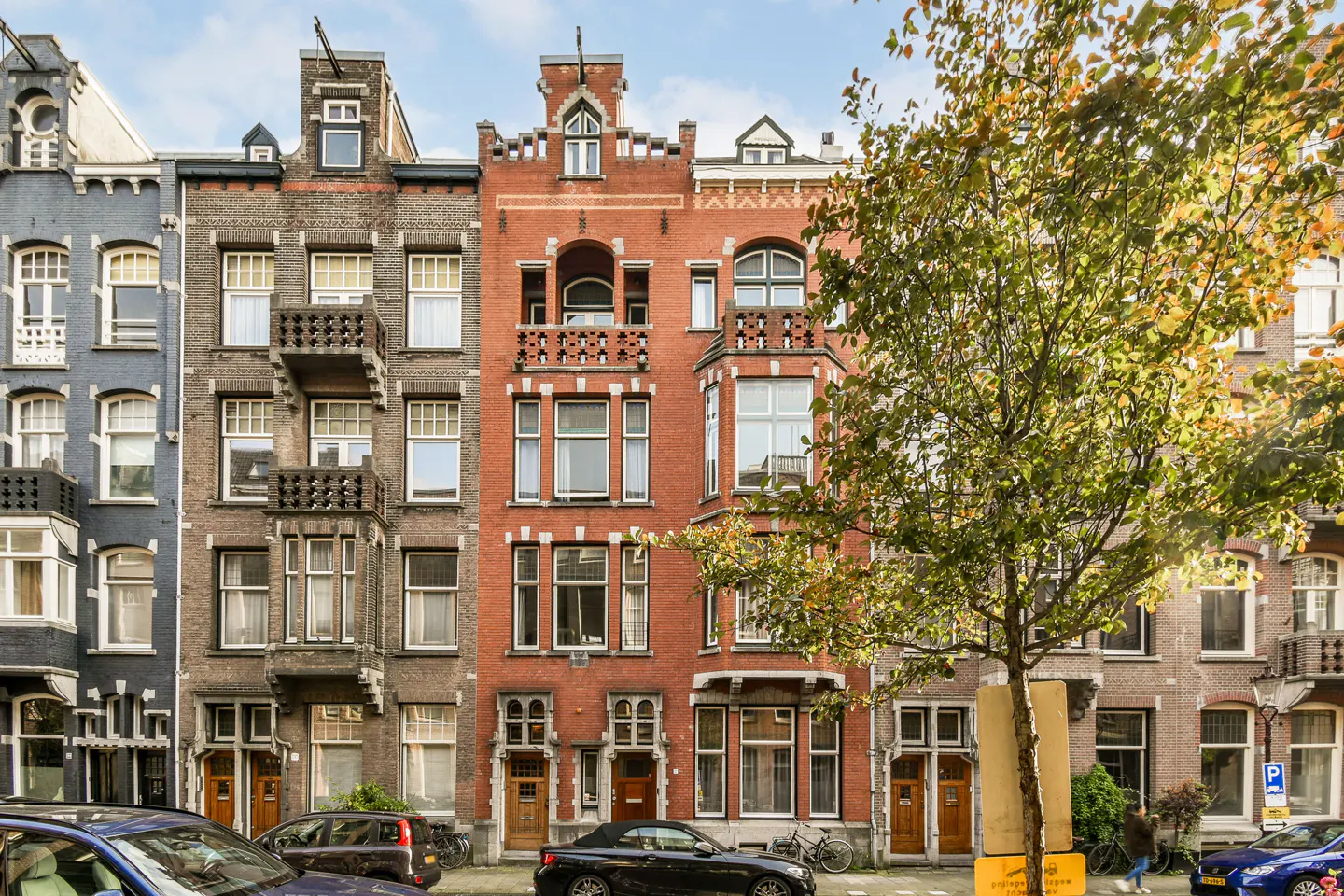 Row of colorful brick townhouses with balconies and large windows, cars parked on the street, and a tree with green and yellow leaves.