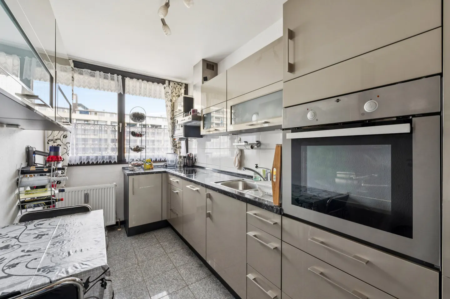 A modern kitchen with beige cabinets, black countertops, and stainless steel appliances. A table sits near a window with sheer curtains.