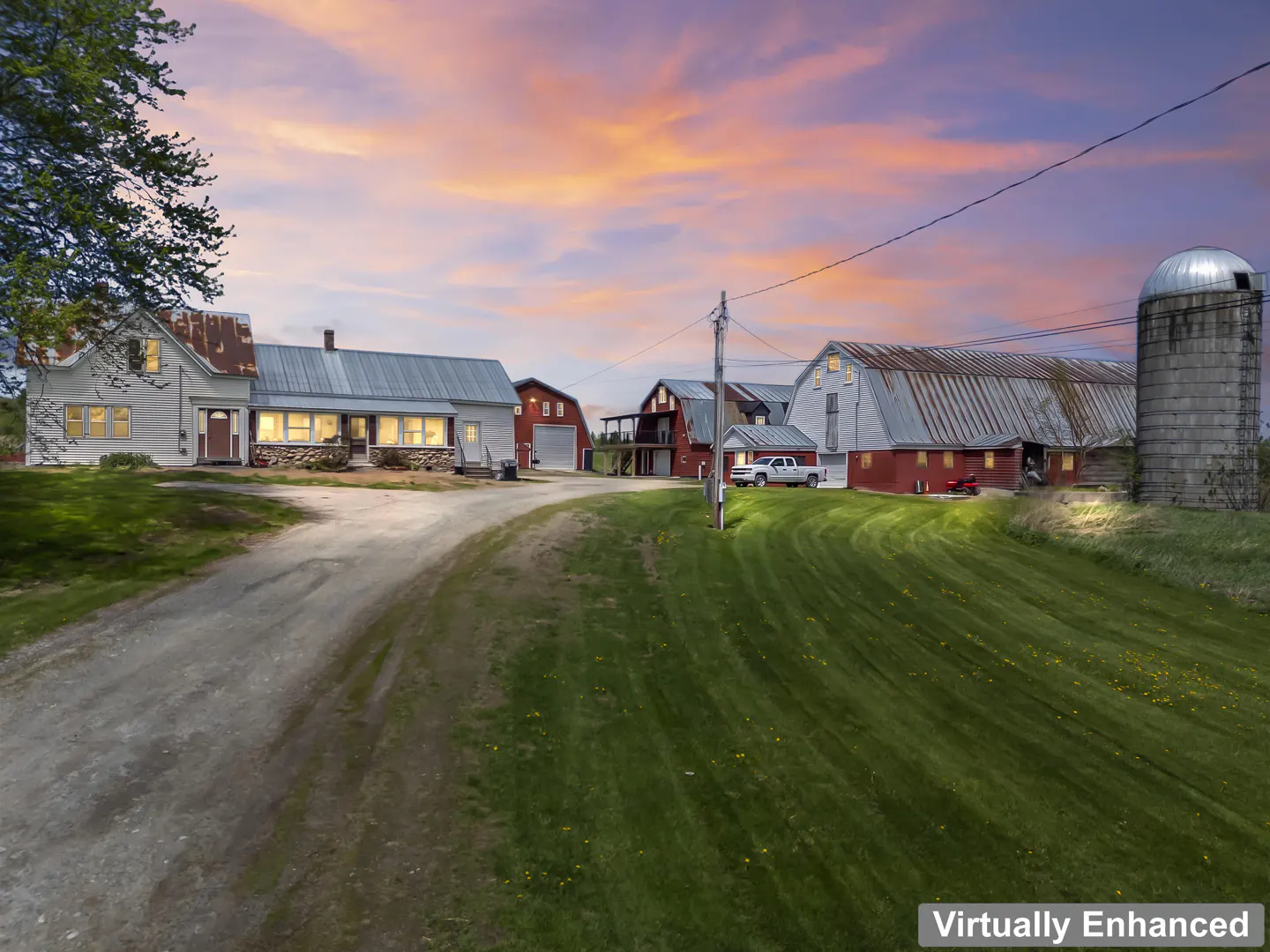 A rural farm with a white house, red barns, and a silo under a colorful sunset sky.
