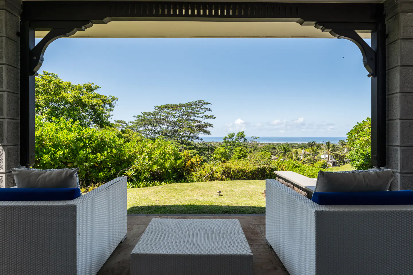 View from a covered patio with white wicker furniture, overlooking a green lawn, trees, and the ocean under a blue sky.