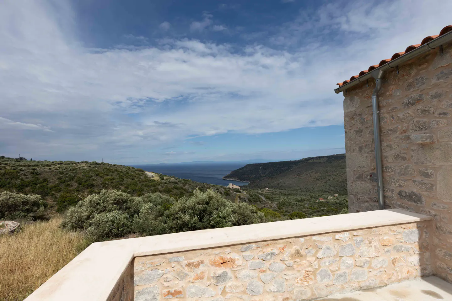 Stone balcony with ocean view. Rolling green hills meet the blue water under a partly cloudy sky. Building with red tile roof on the right.