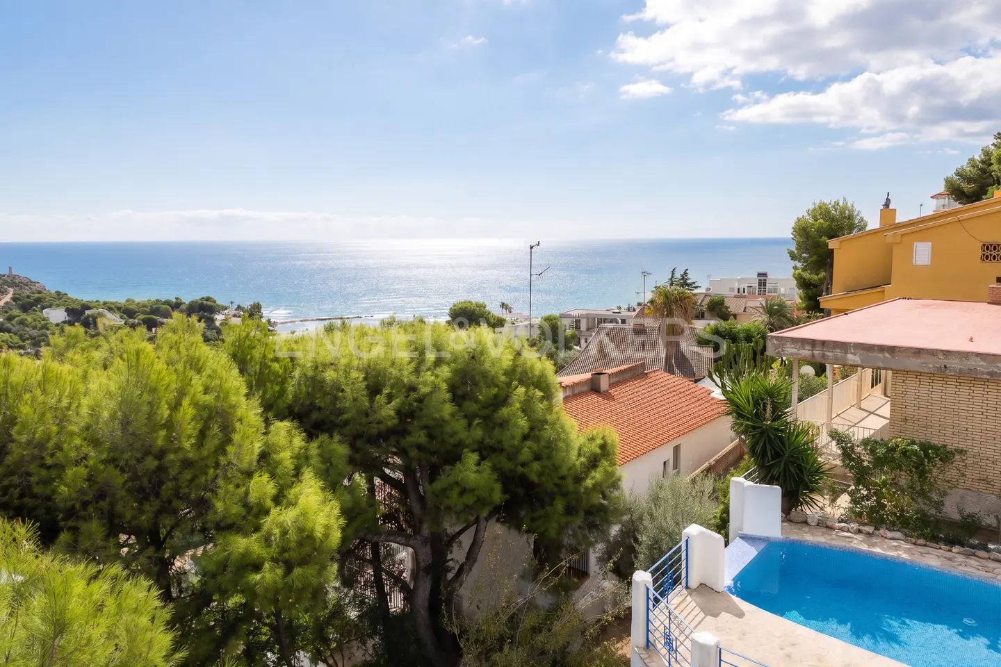 View of a blue pool, trees, and houses overlooking the ocean under a blue sky with clouds.