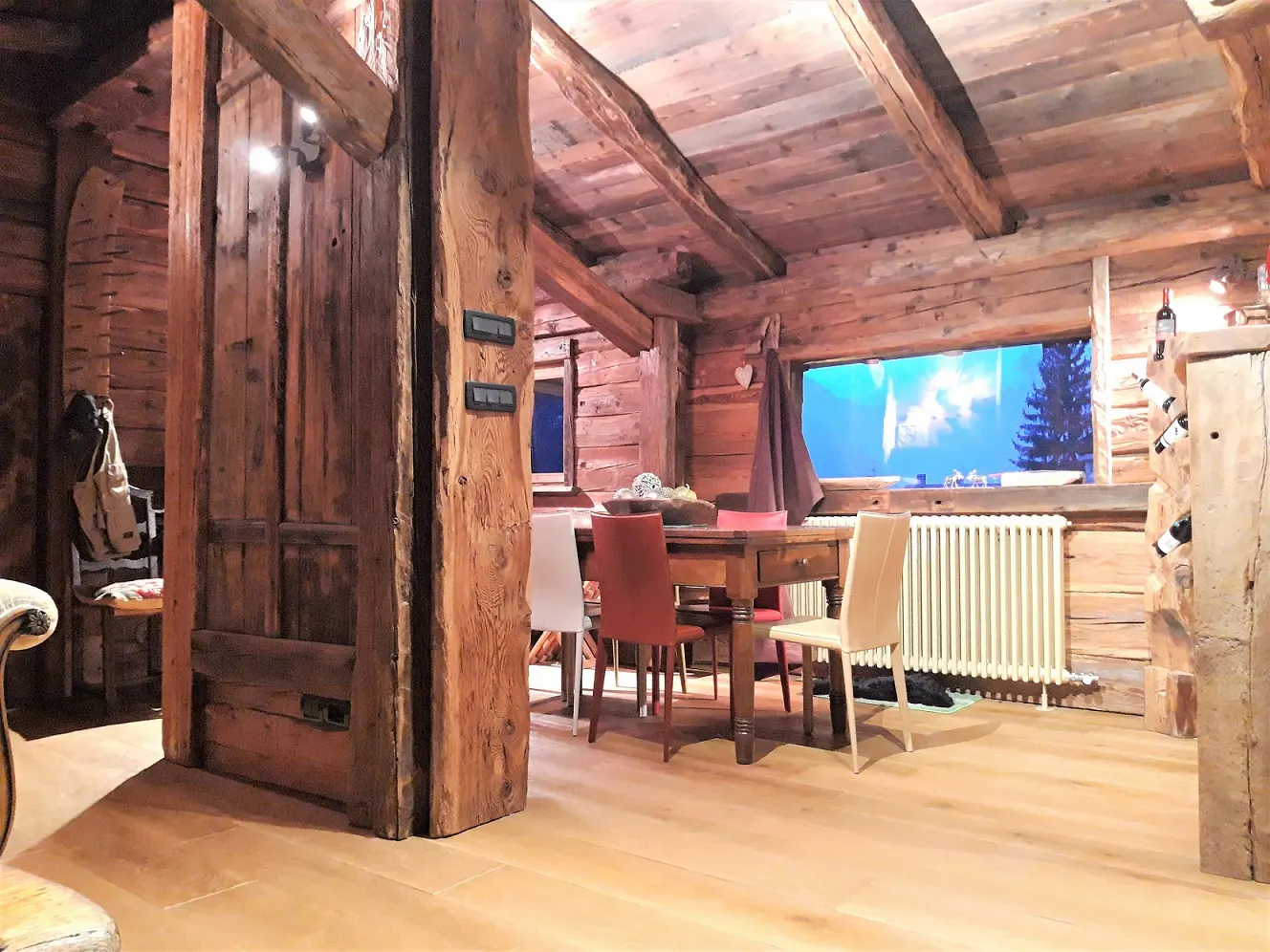 Rustic dining room with wooden walls, ceiling, and floor. A wooden table with chairs sits near a window with a view of a snowy landscape.