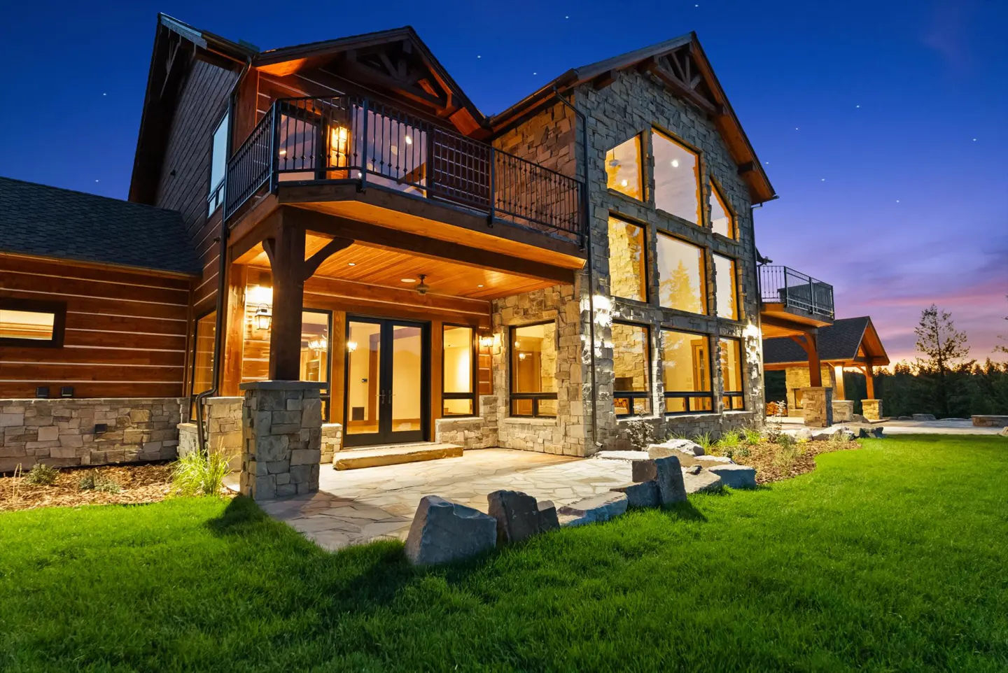 Two-story home with stone and wood exterior, large windows, and balconies under a twilight sky. Green lawn in the foreground.