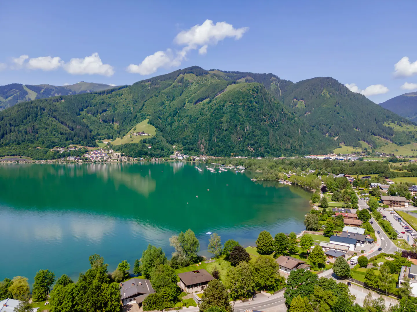 Aerial view of a green lake surrounded by lush green mountains and houses under a blue sky.