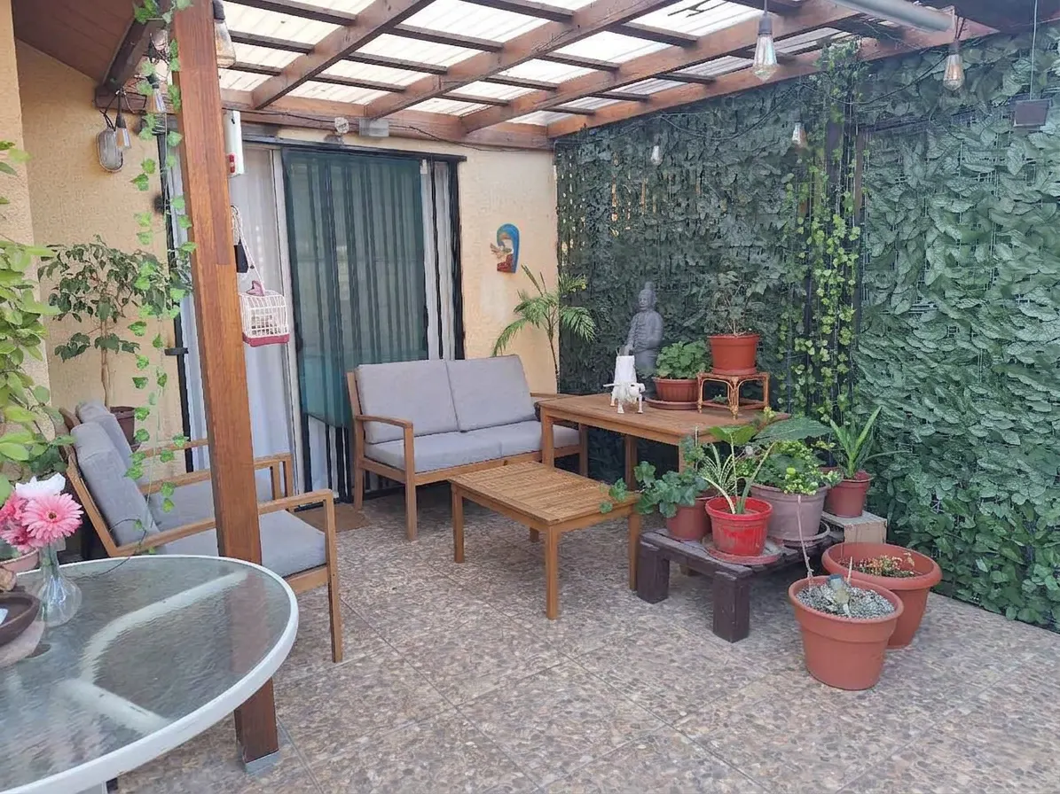 Outdoor patio with gray cushioned seating, wooden tables, and potted plants against a green leafy wall. Overhead is a translucent roof.
